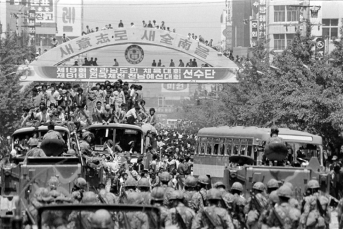 A group demonstrators aboard and atop of buses facing Korean national guard. In the background more protestors are in the street and standing on an arched construction above the street. Black and white photo.