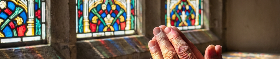 Hands clasped in prayer beside a stained-glass window with colorful light patterns on the skin.