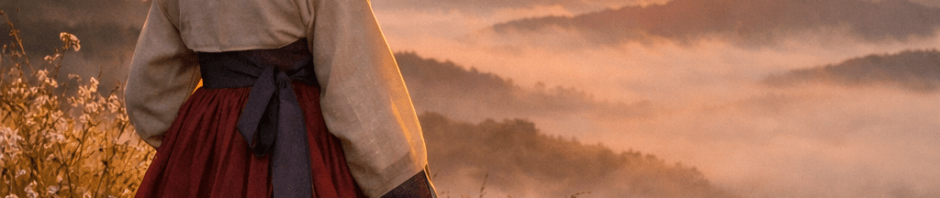Woman wearing traditional Korean hanbok standing near a wooden fence watching sunrise over misty mountains