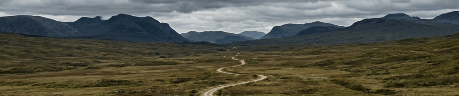 Cyclist on winding dirt trail through moorland with distant mountains and overcast sky